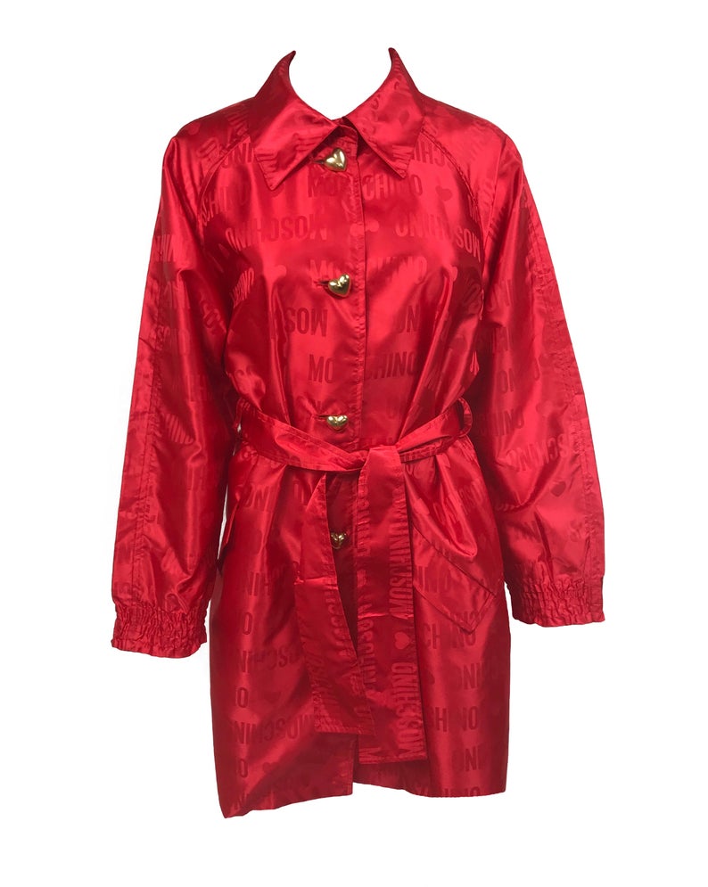 FRUIT Vintage red Moschino Cheap & Chic logo raincoat dating to the 1990s. It features large Moschino logo print all over, heart shaped button closure, front pockets and tie belt.