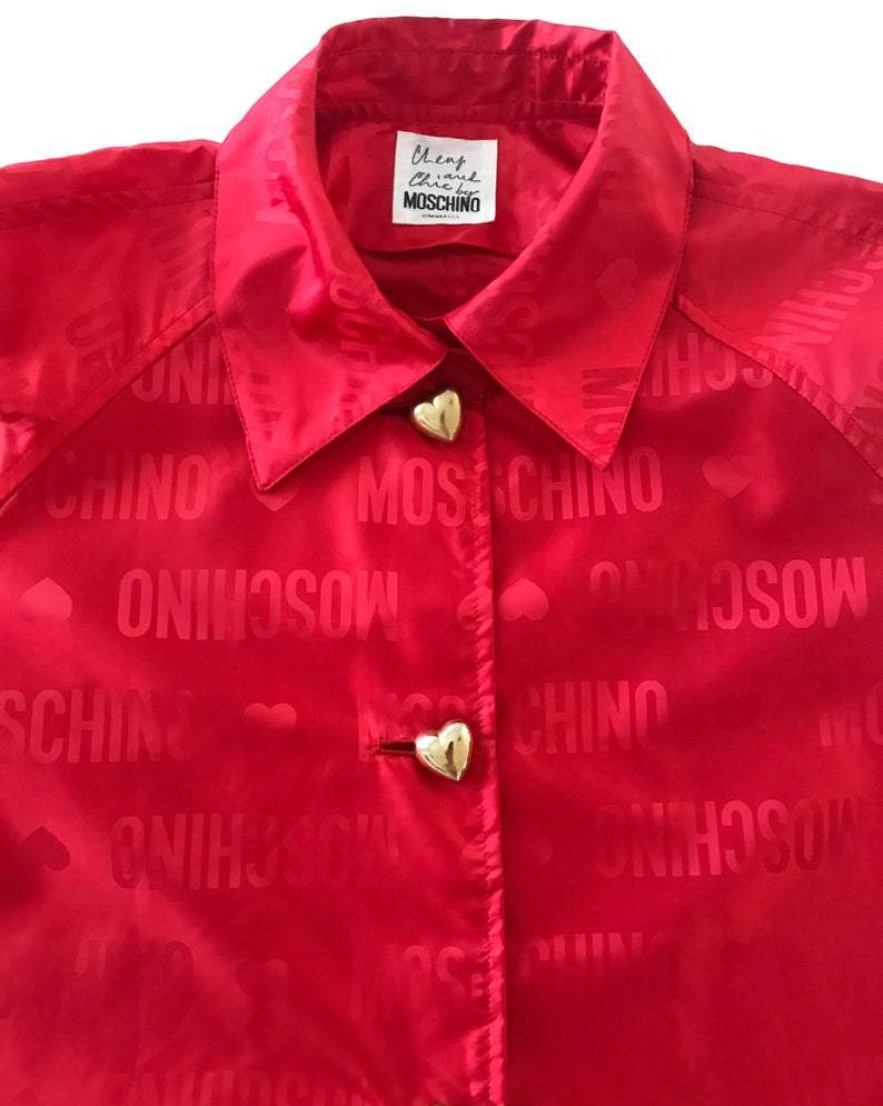 FRUIT Vintage red Moschino Cheap & Chic logo raincoat dating to the 1990s. It features large Moschino logo print all over, heart shaped button closure, front pockets and tie belt.