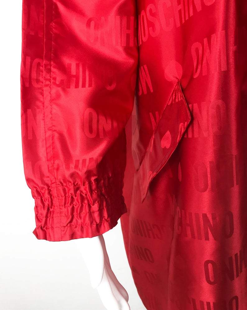 FRUIT Vintage red Moschino Cheap & Chic logo raincoat dating to the 1990s. It features large Moschino logo print all over, heart shaped button closure, front pockets and tie belt.