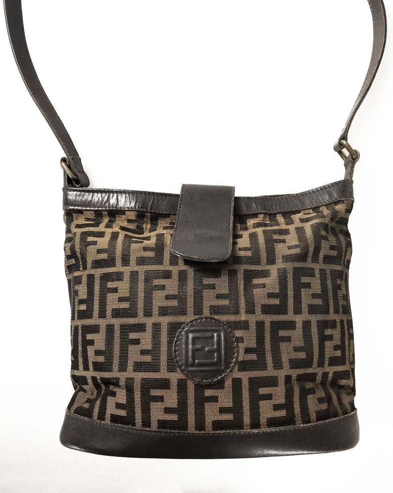 FRUIT Vintage Fendi Zucca cross body bucket bag dating to the 1980s. It features the classic Fendi Zucca monogram canvas, front embossed logo, top push button closure and long adjustable cross body strap.
