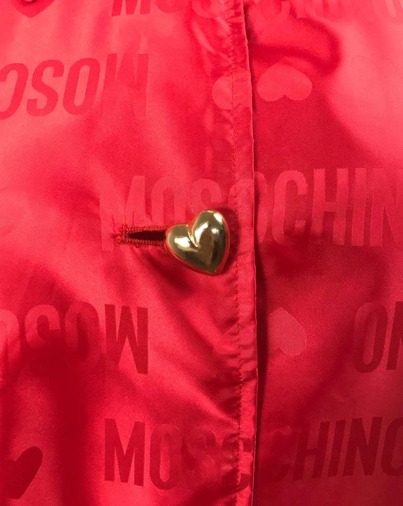FRUIT Vintage red Moschino Cheap & Chic logo raincoat dating to the 1990s. It features large Moschino logo print all over, heart shaped button closure, front pockets and tie belt.